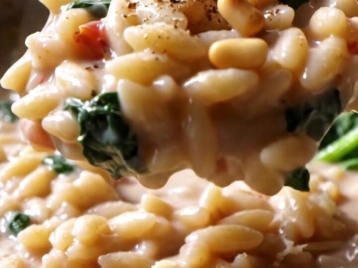 A close-up of creamy Tuscan orzo in a stainless steel pan, topped with pine nuts and cracked pepper.