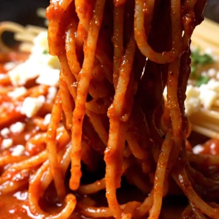 A close-up of crispy, charred spaghetti in a black cast-iron skillet with a red tomato glaze and fresh stracciatella.