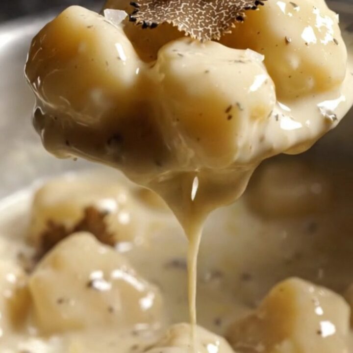 A close-up of pillowy gnocchi coated in a glossy, rich white sauce with visible black truffle shavings.