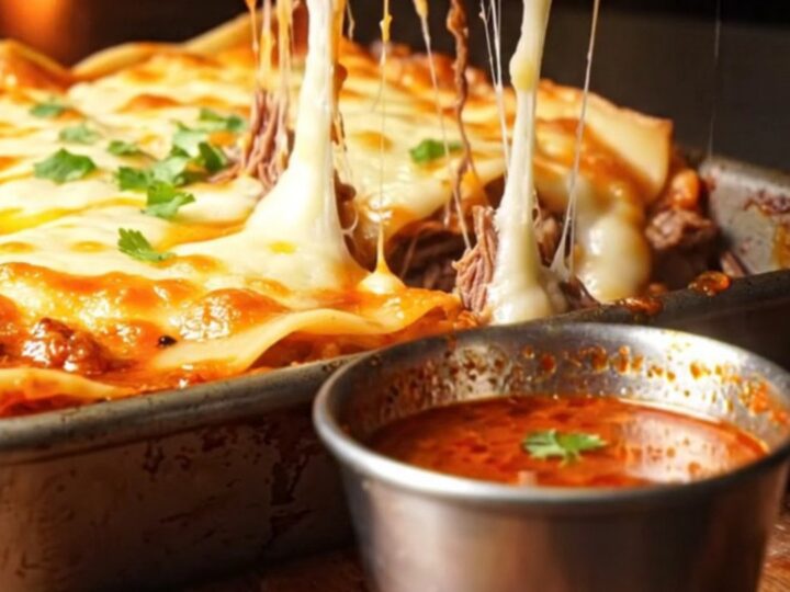 A slice of Quesabirria Lasagna being lifted from a baking dish, showing layers of beef and melted cheese.