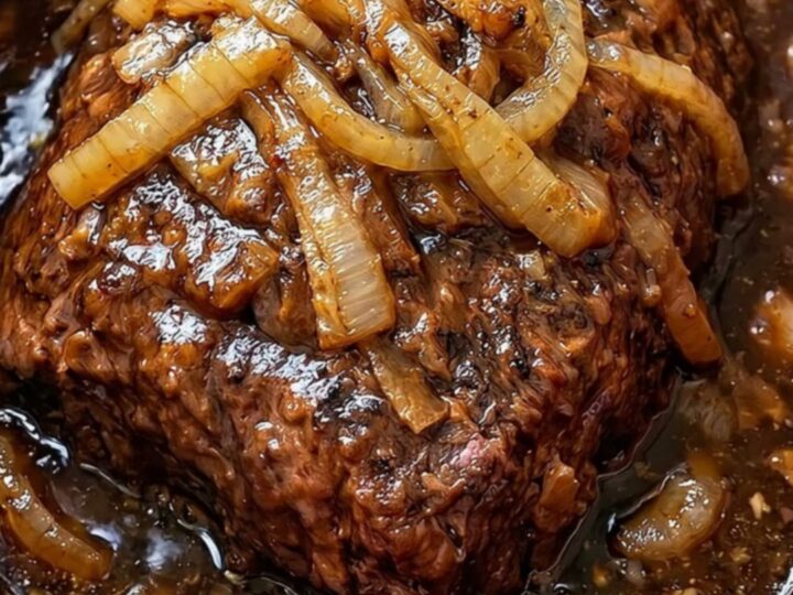 A close-up view of tender shredded beef covered in rich gravy and caramelized onions inside a red slow cooker.