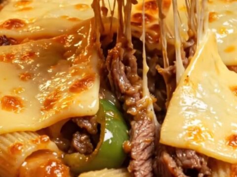 A close-up of a baked rigatoni dish topped with blistered, golden-brown provolone cheese and shaved steak.