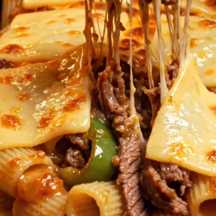 A close-up of a baked rigatoni dish topped with blistered, golden-brown provolone cheese and shaved steak.