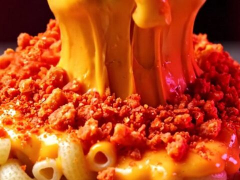 A close-up shot of creamy, molten queso mac and cheese topped with a vibrant, shattered layer of bright red spicy corn chips.