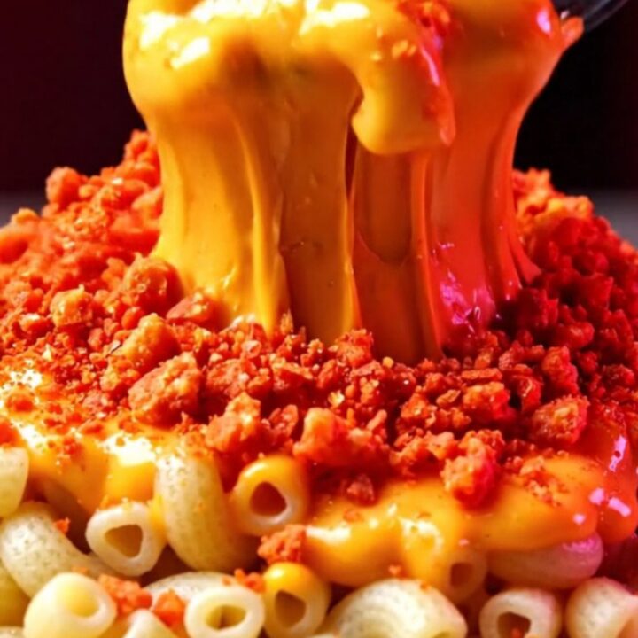 A close-up shot of creamy, molten queso mac and cheese topped with a vibrant, shattered layer of bright red spicy corn chips.