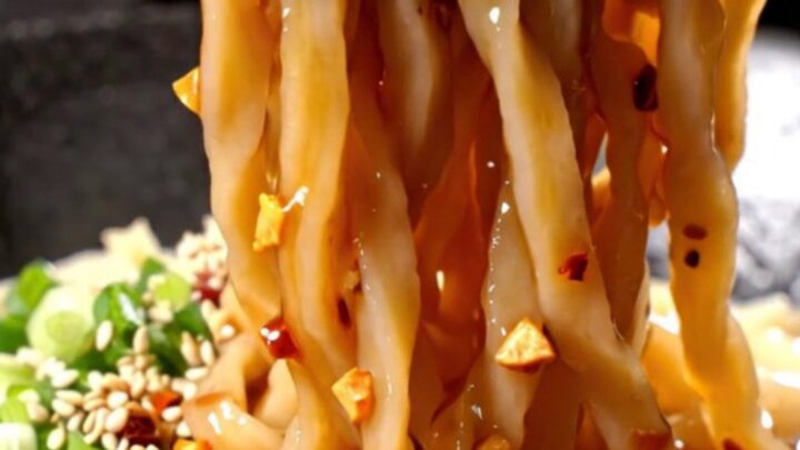 A bowl of thick udon noodles coated in a vibrant, glossy red chili oil sauce with visible garlic and scallions.