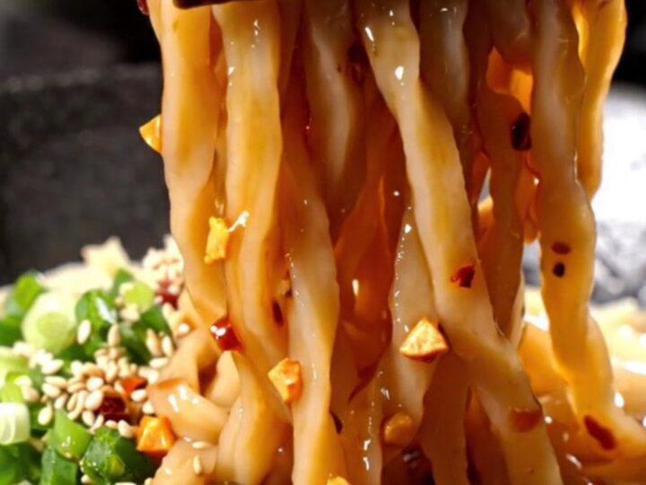 A bowl of thick udon noodles coated in a vibrant, glossy red chili oil sauce with visible garlic and scallions.