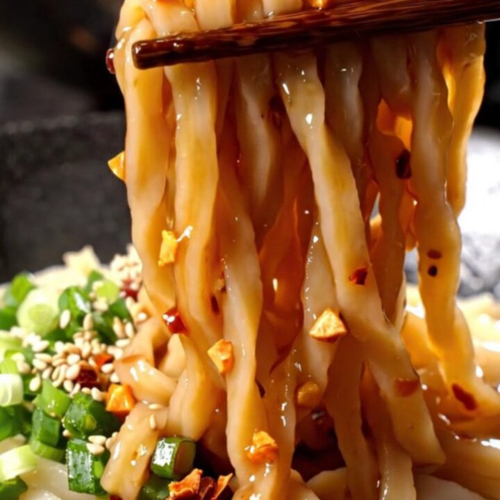 A bowl of thick udon noodles coated in a vibrant, glossy red chili oil sauce with visible garlic and scallions.