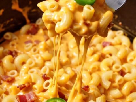 A close-up of a creamy jalapeño popper mac and cheese in a black Dutch oven, topped with crispy bacon and green jalapeños.