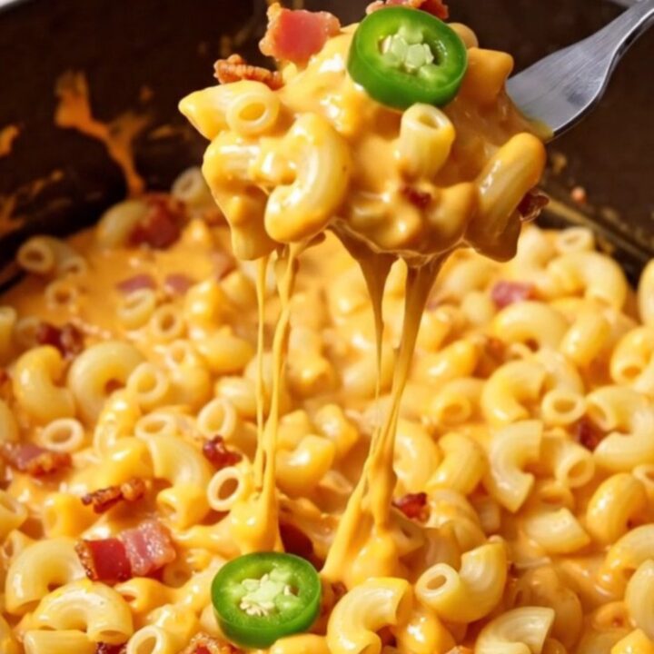 A close-up of a creamy jalapeño popper mac and cheese in a black Dutch oven, topped with crispy bacon and green jalapeños.