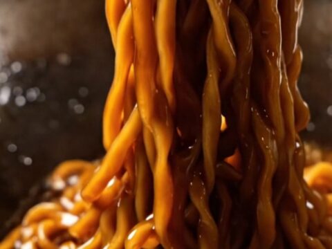 A close-up of glossy dark wheat noodles topped with crispy golden fried scallions.