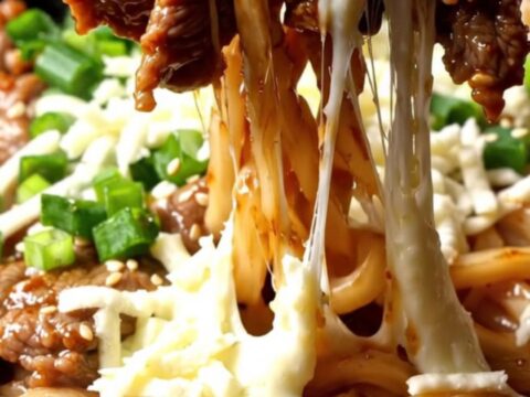A close-up of sizzling beef bulgogi and thick udon noodles topped with melted mozzarella cheese in a hot iron wok.