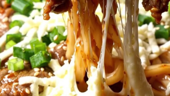 A close-up of sizzling beef bulgogi and thick udon noodles topped with melted mozzarella cheese in a hot iron wok.