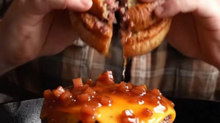 A close-up of a smash burger topped with sticky maple bourbon bacon jam and melted smoked cheddar on a pretzel bun.