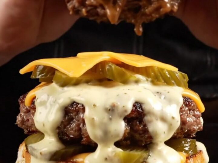 A close-up of a juicy brisket smash burger topped with creamy white sauce and pickles on a toasted bun.