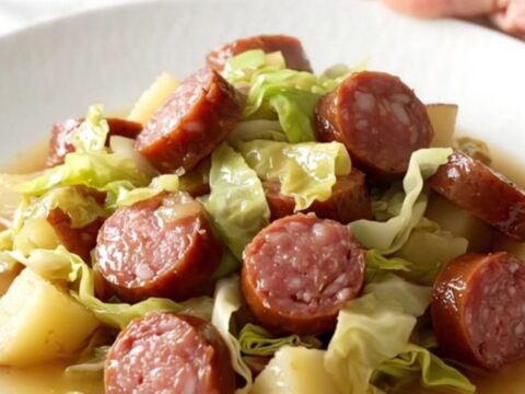 A white ceramic slow cooker filled with sliced smoked sausage, chopped green cabbage, and red potato cubes.