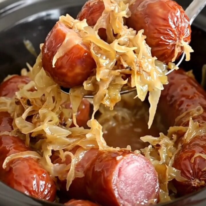 A close-up view of thick chunks of smoked kielbasa served on a bed of golden, soft sauerkraut in a slow cooker.
