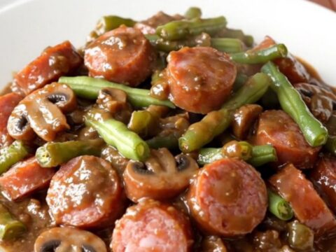 A top-down view of a slow cooker filled with sliced smoked sausage and green beans in a creamy mushroom sauce.