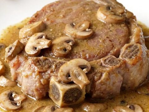 Four raw bone-in pork shoulder steaks arranged in a black oval slow cooker with sliced mushrooms and creamy sauce.