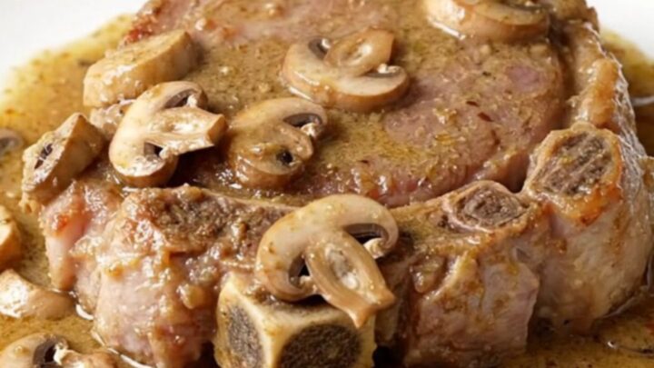Four raw bone-in pork shoulder steaks arranged in a black oval slow cooker with sliced mushrooms and creamy sauce.