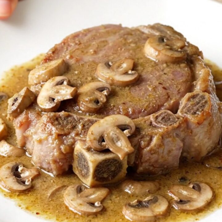 Four raw bone-in pork shoulder steaks arranged in a black oval slow cooker with sliced mushrooms and creamy sauce.