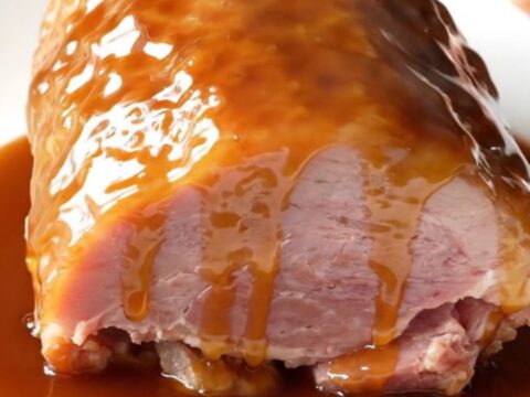 A close-up shot of a glazed, succulent boneless ham on a white plate with a glossy maple brown sugar coating.