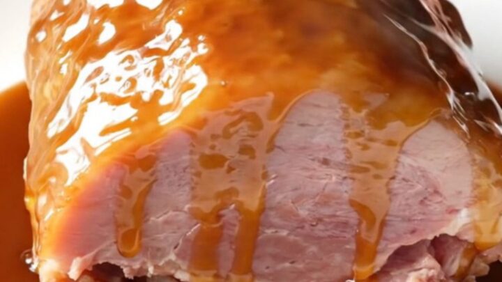 A close-up shot of a glazed, succulent boneless ham on a white plate with a glossy maple brown sugar coating.