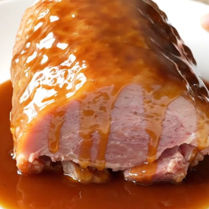 A close-up shot of a glazed, succulent boneless ham on a white plate with a glossy maple brown sugar coating.