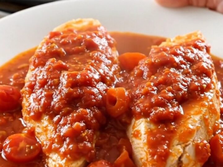 A close-up of tender chicken breasts in a white slow cooker insert, covered in a rich red tomato sauce with cherry tomatoes.