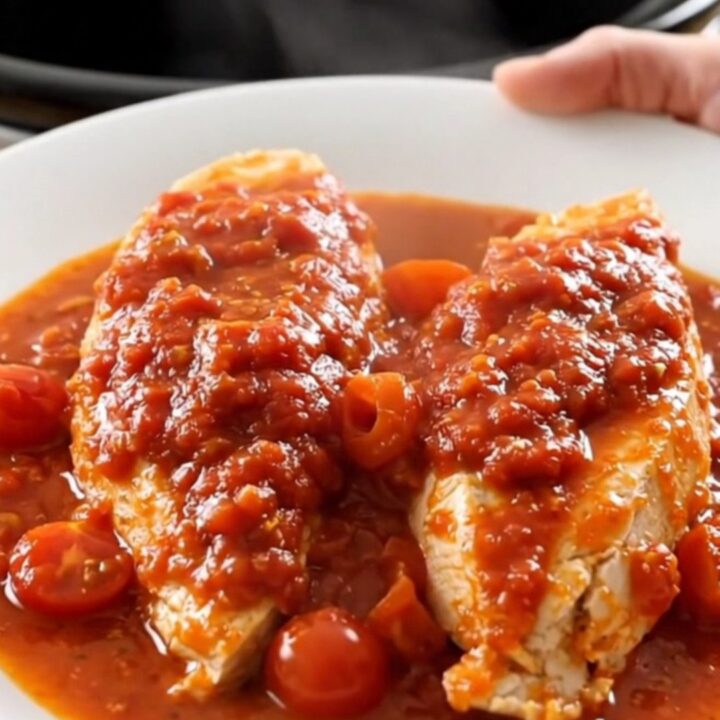 A close-up of tender chicken breasts in a white slow cooker insert, covered in a rich red tomato sauce with cherry tomatoes.