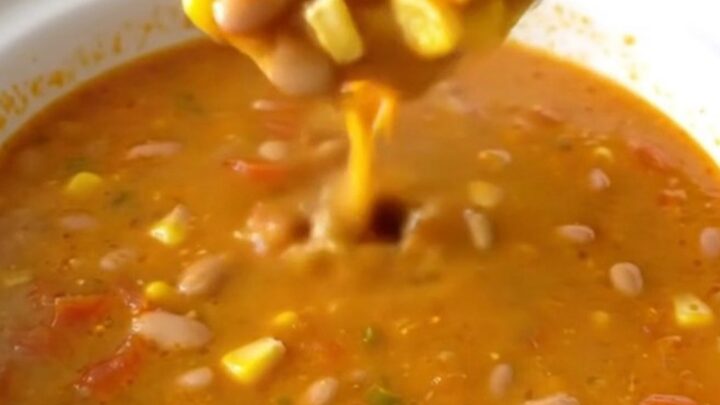 A white shallow bowl filled with creamy orange Santa Fe cheese soup, garnished with pinto beans, corn, and diced tomatoes.