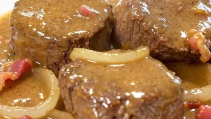 A close-up of tender beef cube steaks smothered in a thick, creamy gravy with bits of crispy bacon and onion slices.