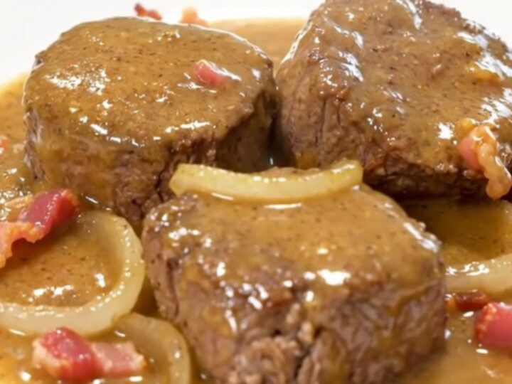A close-up of tender beef cube steaks smothered in a thick, creamy gravy with bits of crispy bacon and onion slices.