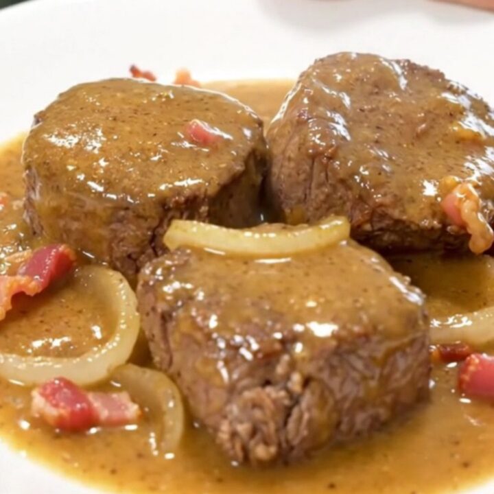 A close-up of tender beef cube steaks smothered in a thick, creamy gravy with bits of crispy bacon and onion slices.