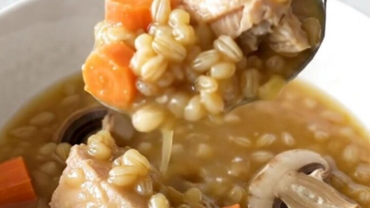 A steaming bowl of chicken and barley stew featuring chunks of chicken, carrot coins, and mushrooms in a rich broth.