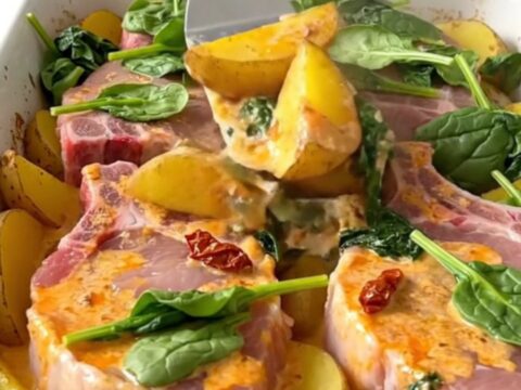 A ceramic baking dish filled with golden pork chops, potato wedges, and a creamy sun-dried tomato sauce with spinach.