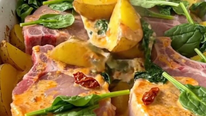 A ceramic baking dish filled with golden pork chops, potato wedges, and a creamy sun-dried tomato sauce with spinach.
