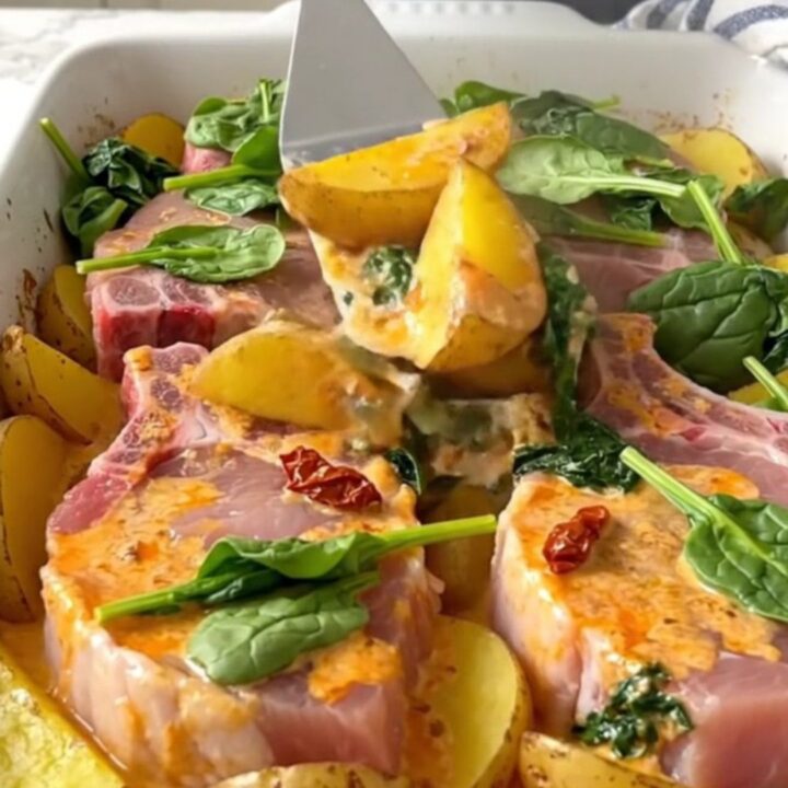 A ceramic baking dish filled with golden pork chops, potato wedges, and a creamy sun-dried tomato sauce with spinach.