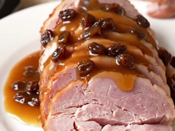 A spiral-sliced ham sitting in a slow cooker, topped with raisins, brown sugar, and pineapple juice.