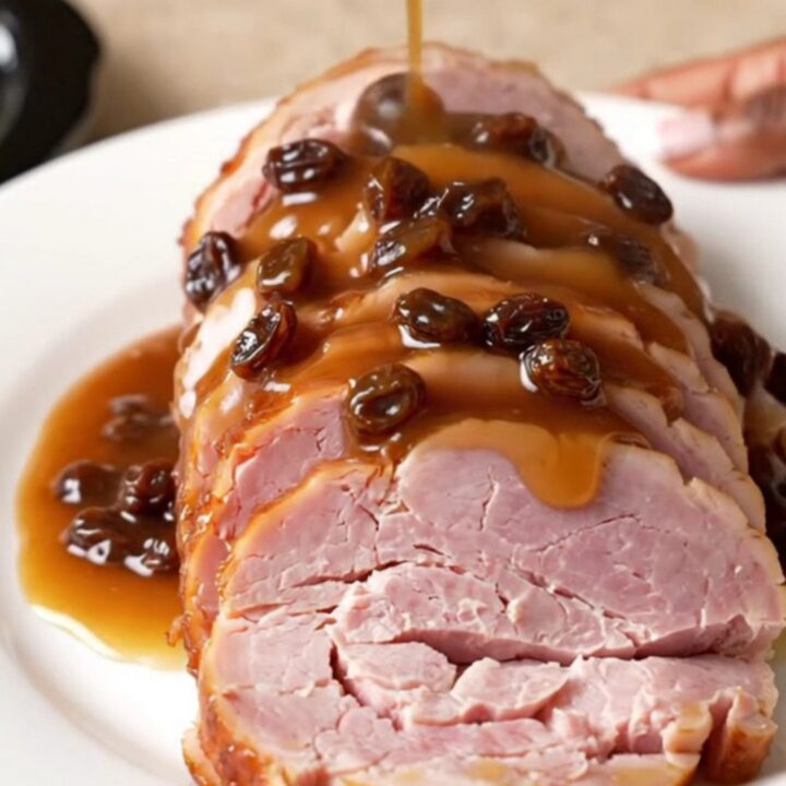 A spiral-sliced ham sitting in a slow cooker, topped with raisins, brown sugar, and pineapple juice.