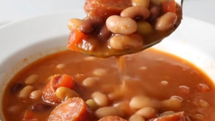 A close-up of a slow cooker filled with a hearty fifteen-bean soup featuring sliced smoked sausage and a rich tomato broth.