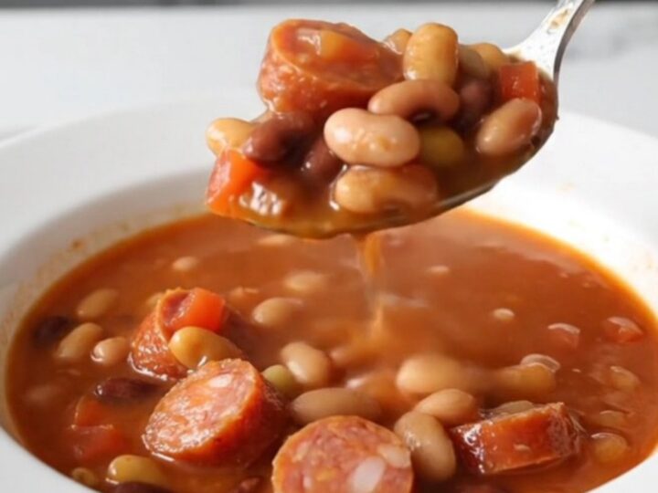 A close-up of a slow cooker filled with a hearty fifteen-bean soup featuring sliced smoked sausage and a rich tomato broth.