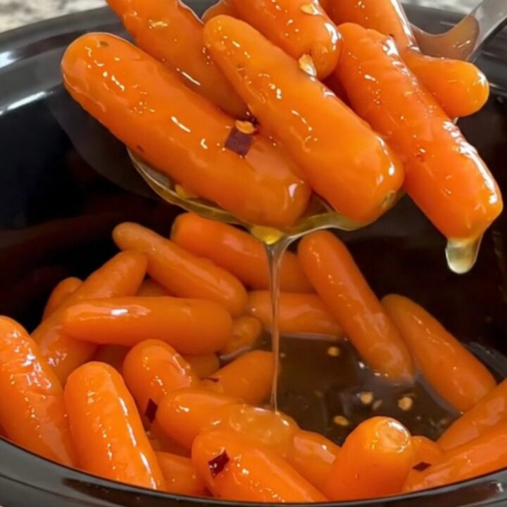 A top-down view of cooked baby carrots in a black slow cooker, glazed with a shiny honey butter sauce.