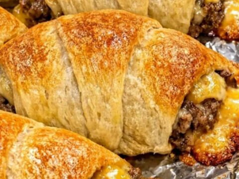 A row of golden-brown cheesy beef crescent rolls arranged on crinkled aluminum foil.