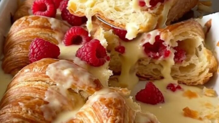 A golden, puffed raspberry cream cheese croissant bake in a white rectangular dish on a granite surface.