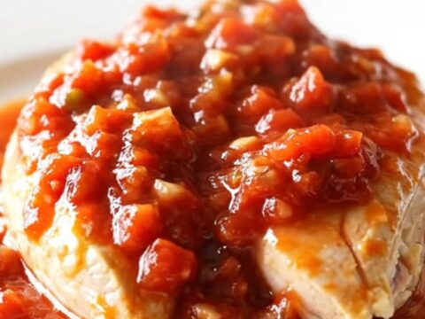 A close-up of slow-cooked chicken breasts smothered in a rich, chunky tomato salsa sauce on a white plate.