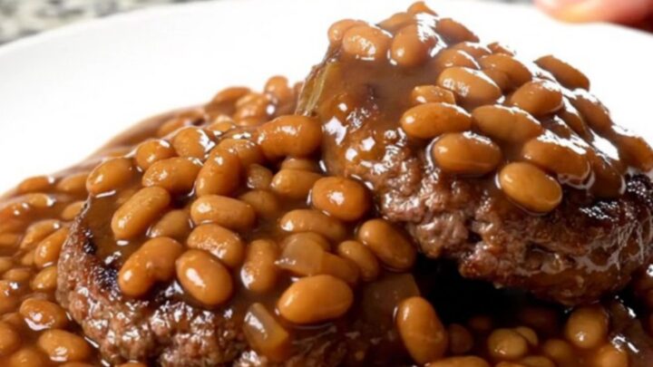 A close-up of tender burger steaks simmered in a rich sauce of baked beans, onions, and green peppers in a slow cooker.
