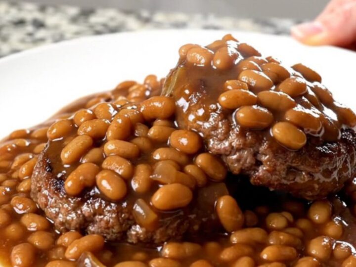 A close-up of tender burger steaks simmered in a rich sauce of baked beans, onions, and green peppers in a slow cooker.