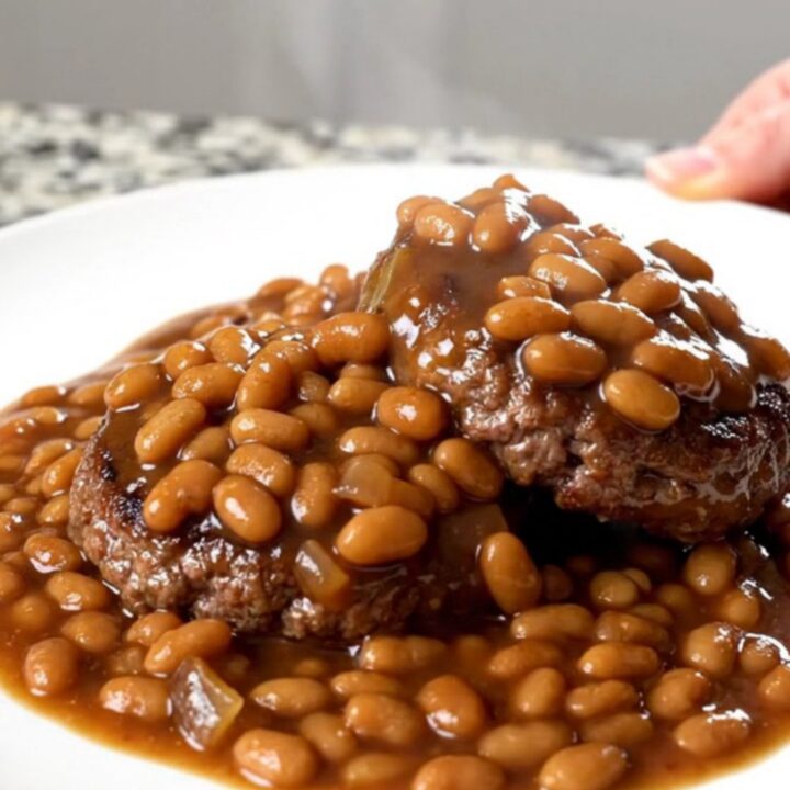 A close-up of tender burger steaks simmered in a rich sauce of baked beans, onions, and green peppers in a slow cooker.