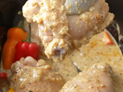 A white slow cooker filled with creamy chicken thighs and vibrant red, orange, and yellow mini peppers.